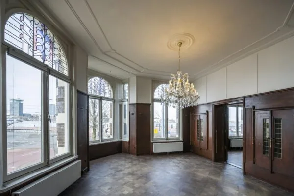 Een statige kamer aan de De Ruijterkade met grote ramen, glas-in-lood details, een houten vloer en een sierlijke kristallen kroonluchter.